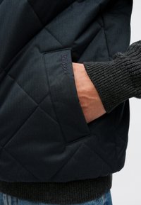 Σκούρο μπουφάν με καπιτονέ ύφασμα και υφασμένο γκρι μανίκι με υφή. Διαθέτει πλαϊνή τσέπη με ανάγλυφο λογότυπο. Το υλικό φαίνεται ανθεκτικό και αδιάβροχο.