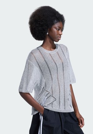 Kvinde med naturlig afro-frisure iført en grå strik top og sorte bukser med hvide striber, stående med den ene hånd i lommen, set fra siden.