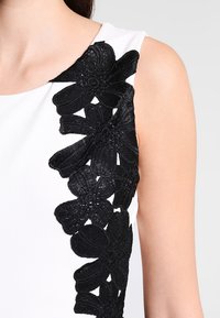 Top blanc avec un motif floral brodé noir le long de l'épaule gauche. Les fleurs sont texturées avec des détails et des motifs complexes.
