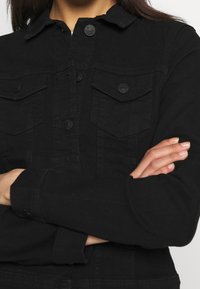 Svart denimjacka med långa ärmar, klassisk krage och två bröstfickor. Har svarta knappar och en strukturerad tyg.