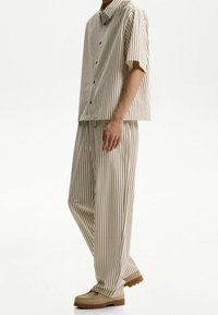 Pruhované tričko s krátkým rukávem a odpovídající kalhoty v béžové barvě s hnědými svislými pruhy. Má volný střih a zapínání na knoflíky. Neformální obuv.