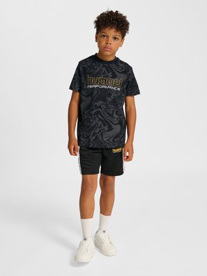 Dreng stående iført sort mønstret "hummel performance" t-shirt, sorte shorts, hvide strømper og hvide sneakers mod ensfarvet baggrund.