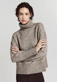 Pull en maille texturée couleur beige à col roulé. Il présente des poignets et un ourlet côtelés. Le modèle se tient debout, les bras croisés, portant une bague en argent.