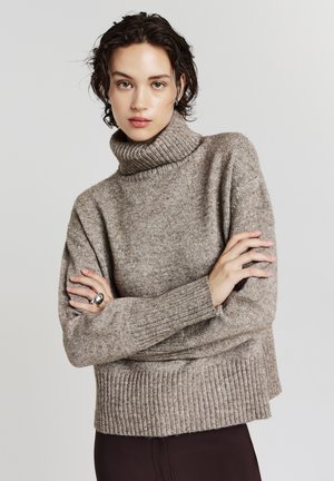 Pullover beige a collo alto in un maglione a trama. Presenta polsini e orlo a costine. Il modello è in piedi con le braccia incrociate, indossando un anello d'argento.