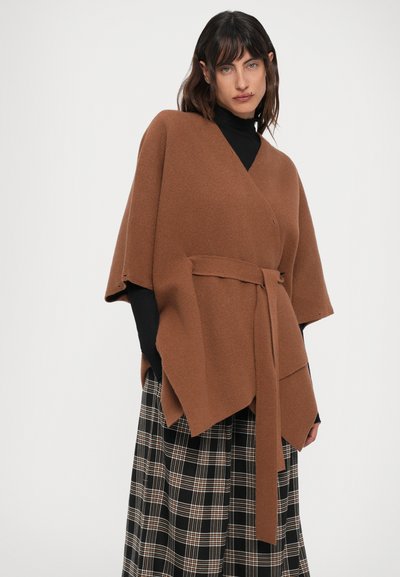 Mujer vistiendo un poncho de lana marrón con cinturón sobre un suéter de cuello alto negro y una falda a cuadros en blanco y negro, de pie contra un fondo blanco.