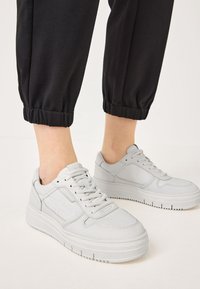 Light gray sneakers with a smooth leather finish, rounded toe, perforated detailing, and a thick, textured sole. Black elastic cuffed pants visible.