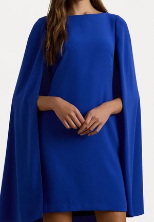 Frau in einem leuchtend blauen Umhangkleid mit langen, fließenden Ärmeln, die Hände vor dem Bauch kontrolliert zusammengelegt, vor einem schlichten Hintergrund.