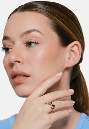 Frau mit heller Haut und zurückgebundenem Haar berührt ihr Gesicht und trägt einen goldenen Schmetterlingsring an ihrer linken Hand mit lackierten Nägeln.