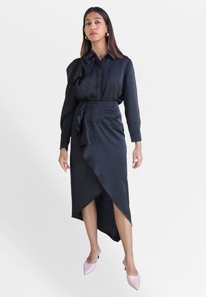 Blouse en satin noir avec un détail en nœud, associée à une jupe taille haute au ourlet asymétrique. Des escarpins rose clair à bout pointu apportent un contraste.