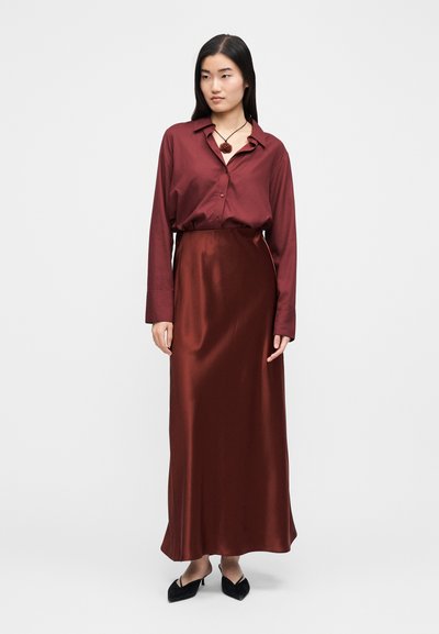 Zestaw z bordową satynową bluzką i długą spódnicą. Bluzka ma guziki i kołnierzyk; spódnica jest gładka o zwiewnej sylwetce. Czarne szpiczaste baleriny.