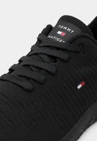 Black sneaker with textured surface, featuring black laces and a branding tag on the tongue. Small logo in red, white, and blue accents.