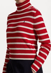 Gros plan sur une personne portant un pull à col roulé rayé rouge et blanc avec des poignets et un ourlet côtelés, associé à un pantalon foncé.