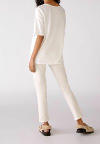 Witte top met korte mouwen en witte strakke broek; model draagt beige slide sandalen met een fuzzy textuur en zilveren hardwaredetails.