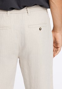 Beige linnen broeken met een subtiele visgraatpatroon, voorzien van achterzakken en een knoopaccent nabij de rechterzak.