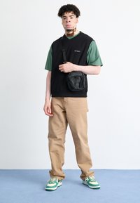 Čierny bezrukávový sveter cez zelené tričko, khaki nohavice a zeleno-biele tenisky. Malá čierna kabelka cez rameno so zipsami. Neutrálne pozadie.