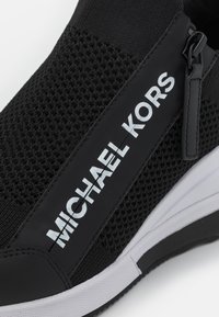 Baskets en mesh noir avec semelle blanche, dotées d'une fermeture éclair latérale et du logo blanc "MICHAEL KORS" sur une bande noire.