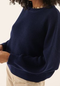 Marinblå stickad tröja med ribbat mönster, rund halsringning och vida ärmar. Kroppen är löst sittande med en nederkant som ligger ovanför midjan.