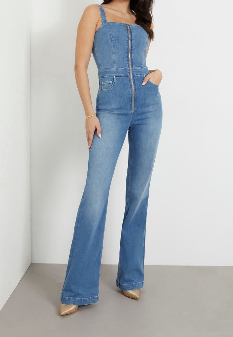 Femme portant une combinaison en denim bleu clair ajustée avec une fermeture éclair à l'avant et des jambes évasées, assortie à des escarpins dorés à bout pointu.