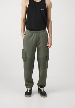 Mand iført sort ærmeløs top og grønne cargo joggerbukser med lynlås ved sidelommerne og sorte sneakers.