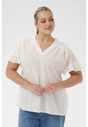 Kobieta o blond włosach związanych nisko w kucyk, ubrana w białą bluzkę z falbanami i niebieskie dżinsy, patrząca na bok z splecionymi rękami.