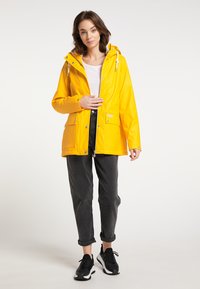 Gele waterdichte jas met een capuchon, zilveren hardware, voorkouwen en een rechte snit, gecombineerd met donkergrijze broek en zwarte sneakers.
