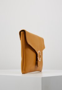 Brązowa skórzana kopertówka z klapką, ozdobiona okrągłym metalowym elementem. Gładka faktura, prostokątny kształt i minimalistyczny design.