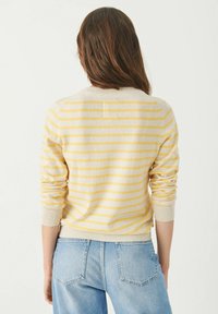 Femme aux longs cheveux bruns portant un pull à rayures crème et jaune et un jean en denim bleu clair, tournée dos au spectateur.