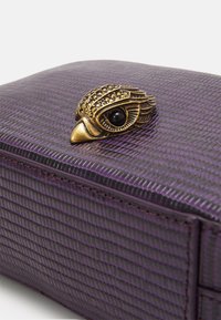 Lila texturerad handväska med ett bronzfärgat fågelformad lås som har ett svart ädelstenöga, vilket ger en unik accent till dess design.