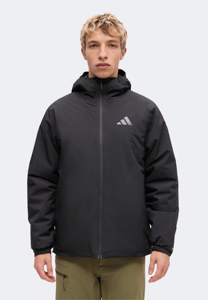Junge Person mit kurzen blonden Haaren, die eine schwarze Kapuzenjacke mit reflektierendem Adidas-Logo und olivgrünen Hosen trägt, schaut nach vorne.