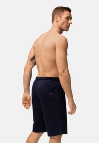 Oberkörperfreier Mann mit kurzen Haaren, der marineblaue Shorts mit elastischem Bund trägt, sich leicht zur Seite dreht und ein Tattoo auf seinem linken Arm zeigt.