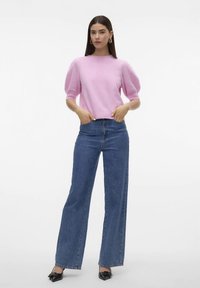 Sieviete, tērpusies rozā puff-sleeve džemperī, augsto jostasvietu platās zilo džinsu biksēs, melnajos norādītā pirksta zābakos un zeltītās auskaros, stāv ar rokām kabatās.