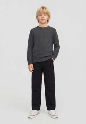 Băiat blond stând cu o mână în buzunar, purtând un pulover gri închis, pantaloni negri și adidași albi pe un fundal simplu, deschis.