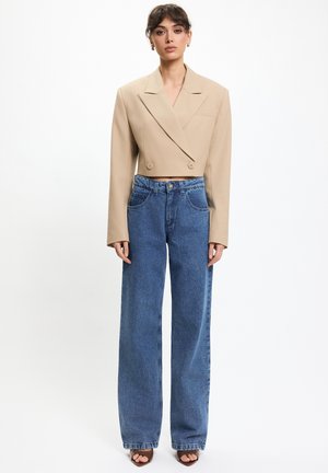 Kobieta ubrana w beżową, krótką, dwurzędową marynarkę, niebieskie, szerokie spodnie z wysokim stanem oraz sandały na obcasie, stojąca na tle białego tła.