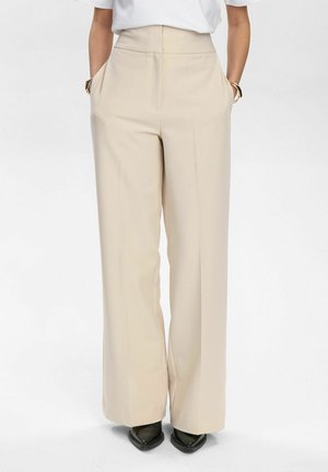 Pantaloni beige a gamba larga con vita alta, dotati di due tasche laterali, tessuto morbido e taglio dritto che termina sopra le scarpe nere.