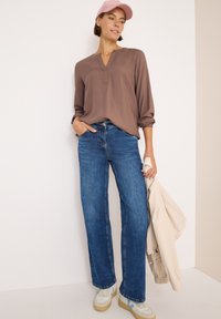 Maglia lunga a maniche con scollo a V di colore marrone intrecciata; jeans a gamba larga blu; sneakers chiare con accenti blu; giacca chiara tenuta in una mano; berretto rosa.