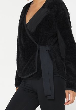 Frau trägt eine schwarze, strukturierte Wickeljacke mit einem Stoffgürtel, der in der Taille gebunden ist, und schwarze, eng anliegende Hosen.