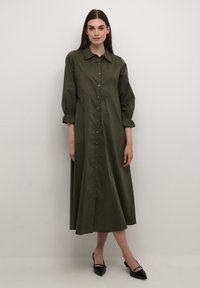 Olive green, long-sleeve button-up dress with a collar, gathered sleeves, and a mid-length skirt, paired with black pointed-toe shoes.