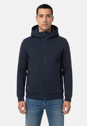 Uomo in piedi che indossa una giacca con cappuccio blu navy con zip e jeans blu su uno sfondo chiaro e uniforme.