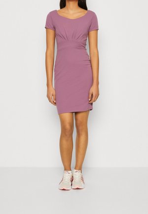 Femme portant une robe mauve à manches courtes et longueur genou, assortie de chaussures de sport blanches et roses, debout devant un fond clair uni.