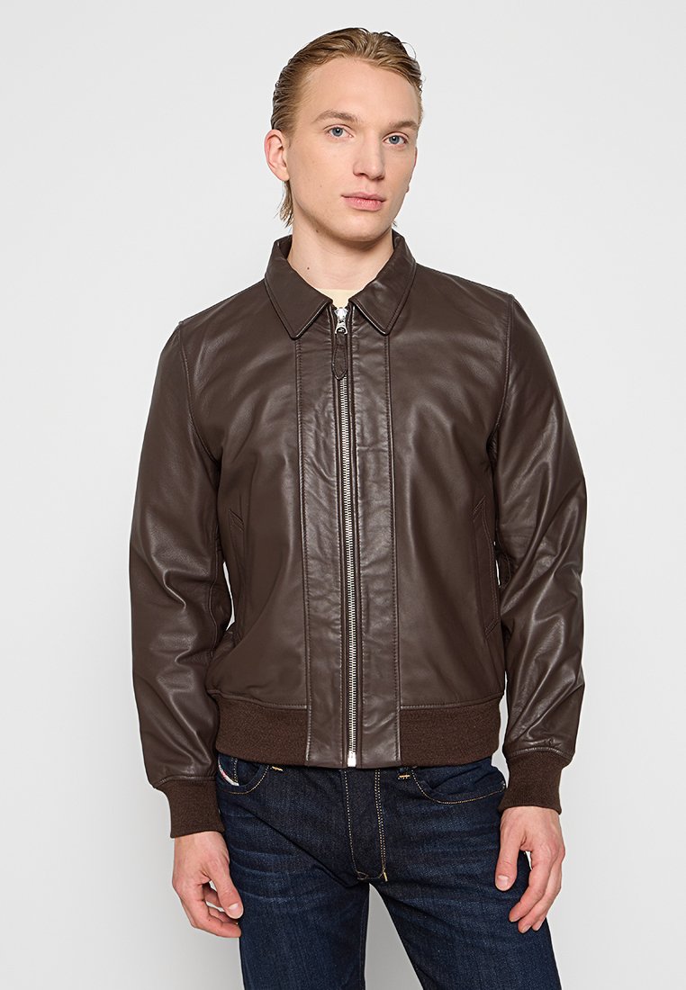 Veste bomber en cuir marron avec fermeture éclair, poignets et col côtelés ; texture lisse et design ajusté ; associée à un jean en denim foncé.