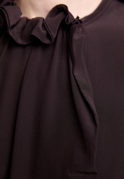 Blusa marrone scuro con scollatura arricciata e dettaglio a occhiello nella parte anteriore. Il tessuto sembra leggero e liscio al tatto.
