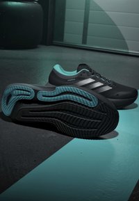 Chaussures de course Adidas noires et bleu sarcelle avec tige Primeknit et semelle à motifs, placées sur un sol sombre avec des accents lumineux bleu sarcelle.