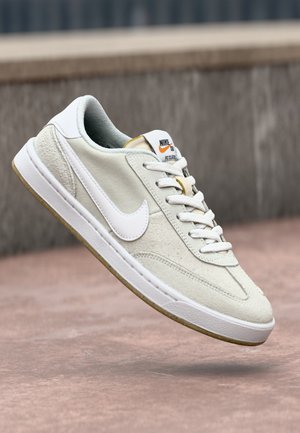 Baskets Nike blanches et beiges avec semelle en gomme et lacets, présentées en angle au-dessus d'une surface en béton et d'un mur gris en arrière-plan.