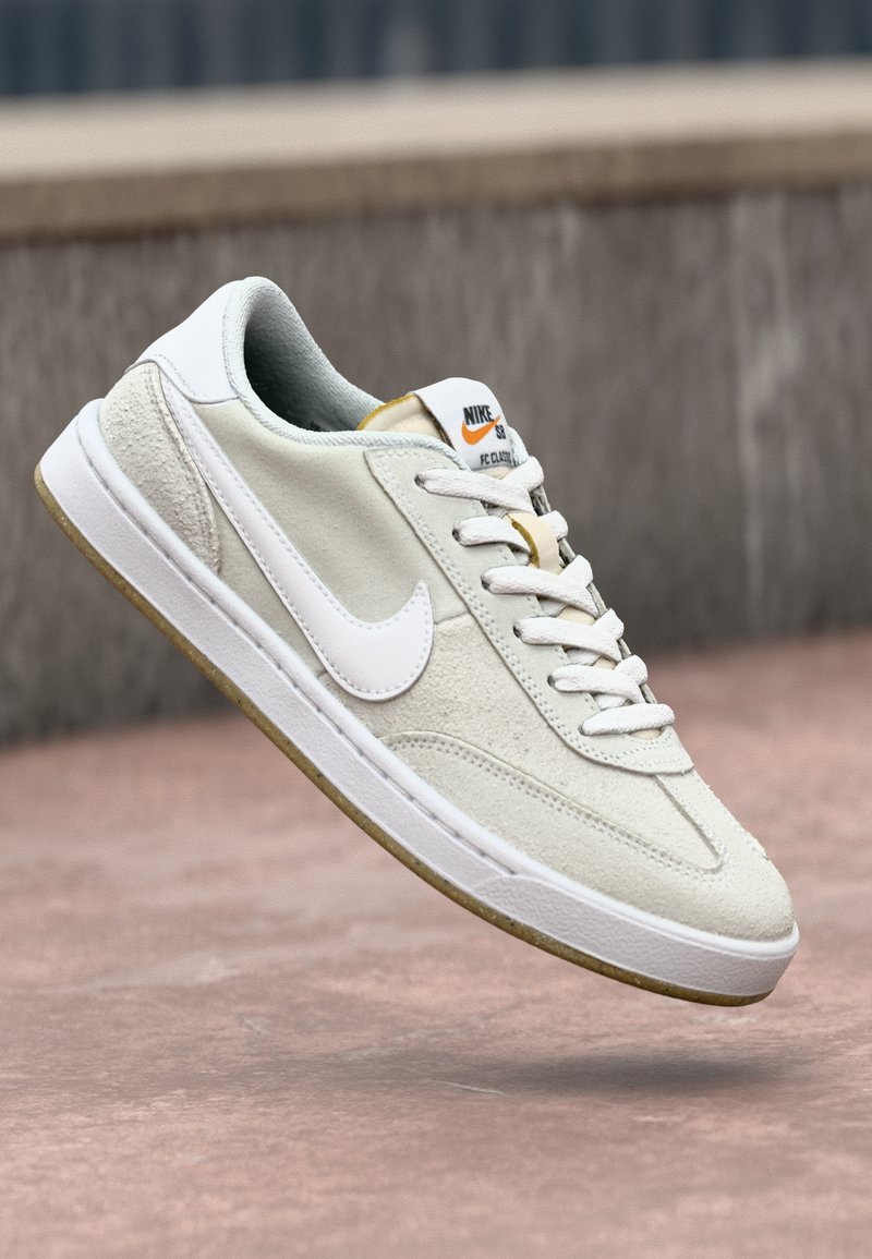 Baskets Nike blanches et beiges avec semelle en gomme et lacets, présentées en angle au-dessus d'une surface en béton et d'un mur gris en arrière-plan.