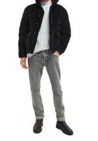 Giacca puffer di un blu navy scuro con cappuccio, caratterizzata da chiusure a scatto e tasche laterali. Indossata sopra un maglione testurizzato blu chiaro, abbinata a jeans grigi e stivali neri.