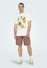 Camiseta de algodón blanca con un gráfico de palmera y el texto "PALM BEACH", combinada con pantalones cortos de cargo marrones y zapatillas ligeras con calcetines blancos.
