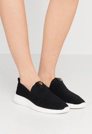 Jambes nues portant des baskets en mesh noires sans lacets avec semelles blanches et petit emblème doré sur le dessus sur fond uni.