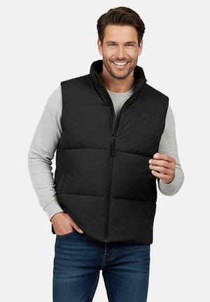Lachende man met een zwart donsvest over een lichtgrijs shirt met lange mouwen en een blauwe spijkerbroek, één hand in de zak, tegen een eenvoudige achtergrond.