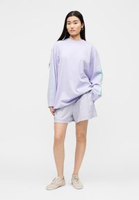 Femme portant une chemise lavande ample à manches longues, un short à carreaux et des baskets beiges, debout avec une main dans la poche sur un fond uni.