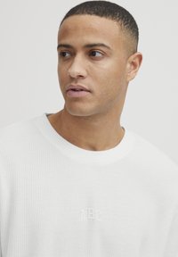 Jeune homme aux cheveux courts portant un pull en tricot blanc texturé, regardant à sa gauche contre un fond clair et uni.